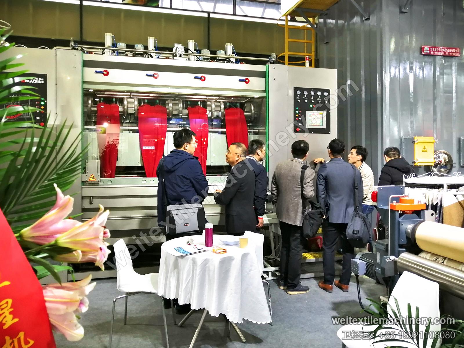 2019 年展台，来宾围在四通道洗缩联合机前，四个通道里挂着红色面料